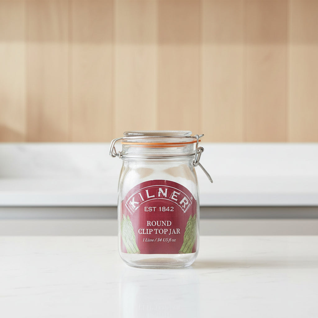 A Kilner round clip top jar made of clear glass with a dark red oval label and metal wire clamp lid, shown on a white background.