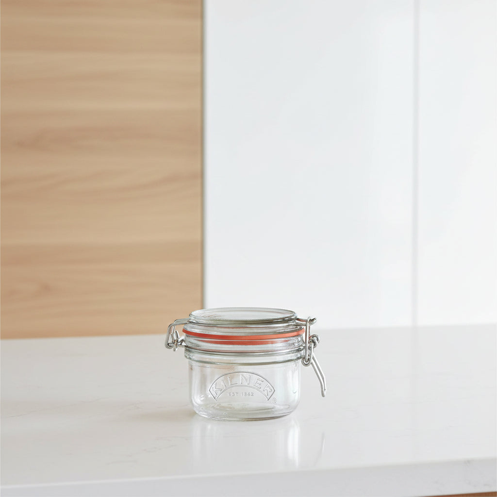 A Kilner glass jar with a metal clamp lid and orange-red rubber gasket, shown on a plain white background.