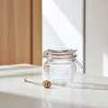 A clear ribbed glass jar with a metal clamp lid and a wooden honey dipper, shown on a plain white background. The jar is embossed with the brand name Kilner.