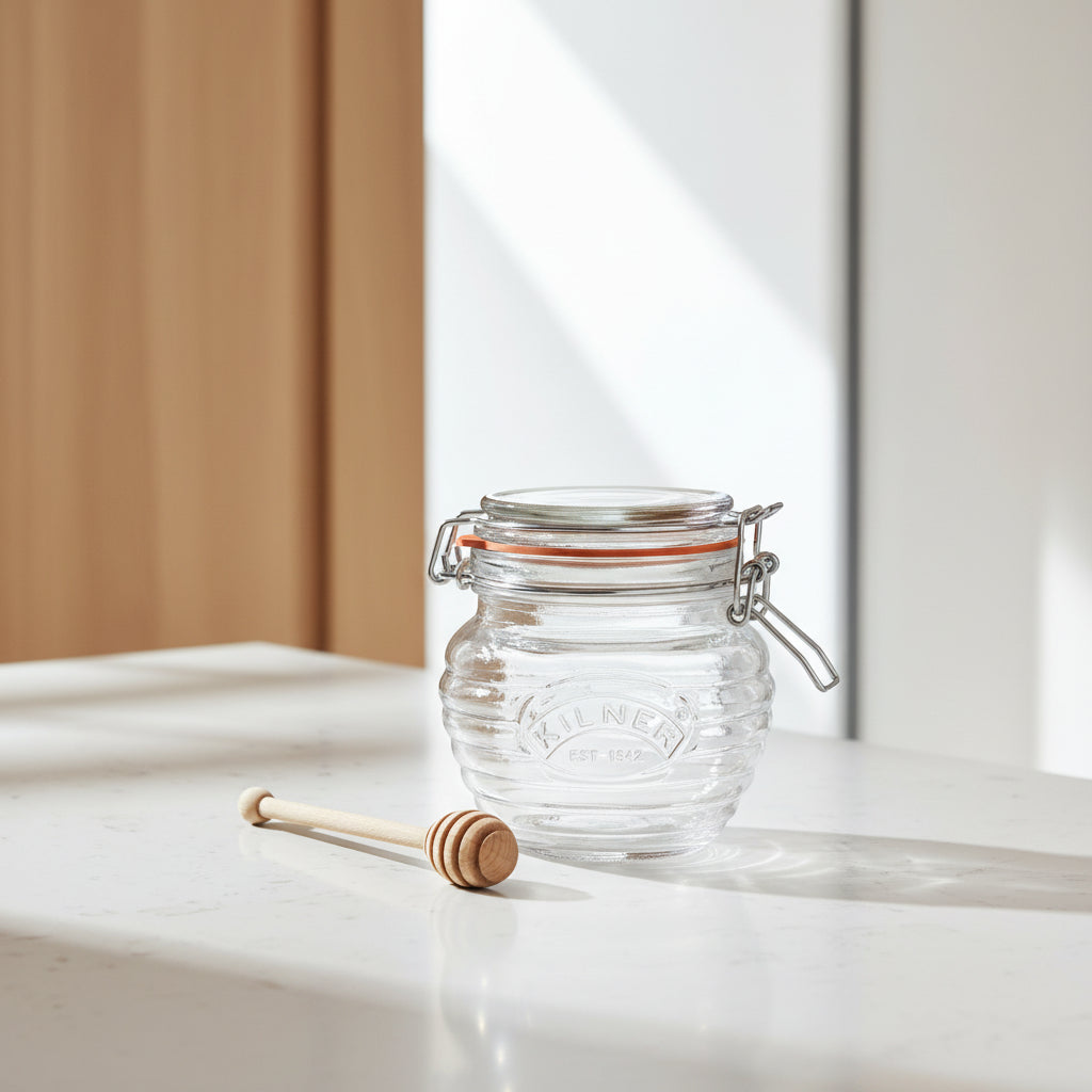 A clear ribbed glass jar with a metal clamp lid and a wooden honey dipper, shown on a plain white background. The jar is embossed with the brand name Kilner.