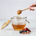 Glass jar of honey with a wooden dipper, surrounded by fruits and popcorn on a wooden surface.
