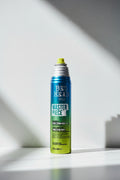 A can of Tigi Bed Head Master Piece hair spray, featuring a metallic blue-to-yellow gradient design with white and green text, set against a dark, moody studio background.