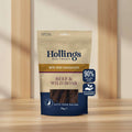 A 70g bag of Hollings Dog Treats Beef & Wild Boar Bite-Size Sausages. The packaging is beige on top and dark navy blue on the bottom, with a clear window showing the dark brown sausage pieces inside.