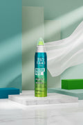 A can of Tigi Bed Head Tige Master Piece styling mousse stands on a purple surface against a bright green and blue geometric backdrop. The can is metallic teal and green with white text, and the right side of the image includes a black-and-white abstract patterned panel.