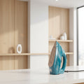 A teal and white Russell Hobbs steam iron stands on an ironing board next to a stack of folded white, gray, and yellow shirts in a softly blurred bedroom setting.