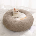 A fluffy round cat bed in light taupe, with a long-haired white and orange cat resting inside, set on a white faux fur rug in front of sheer white curtains.