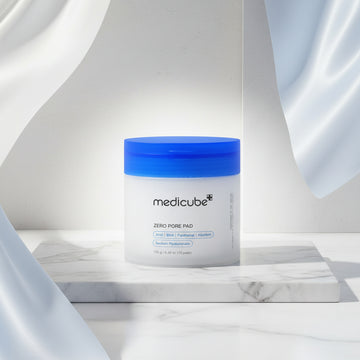 A white jar of medicube+ Zero Pore Pad with a bright blue lid, shown against a white circular background with a faint texture.