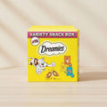 Dreamies Variety Snack Box featuring a bright yellow package with a cartoon cat, snack pieces, and small illustrated snack pouches. The box prominently displays the brand name 'Dreamies' and 'VARIETY SNACK BOX' with 'x12' indicating a 12-pack.