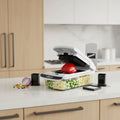A white and black Fullstar vegetable chopper and grater countertop appliance shown open with a red bell pepper inside, alongside inset graphics of different blade attachments and chopped vegetables.