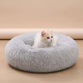 A fluffy round gray pet bed on a white faux-fur rug, with a small white cat sitting inside. A gray sofa leg is visible in the background.