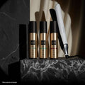Four g/100 haircare bottles in metallic gold and black arranged on a black marble pedestal, with a white and black g/100 hair straightener leaning against them on a dark studio background.