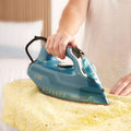 A person ironing a yellow patterned fabric with a Russell Edson motorised steam iron in a blue and teal color scheme, set against a softly blurred bedroom background.
