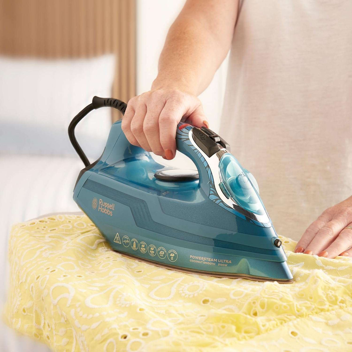 A person ironing a yellow patterned fabric with a Russell Edson motorised steam iron in a blue and teal color scheme, set against a softly blurred bedroom background.
