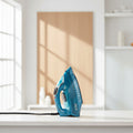 A blue and teal Russell Hobbs steam iron with a transparent water tank, standing on a white countertop with a black power cord. The background features a white brick wall and a wooden cutting board with kitchen items.
