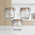 Three clear Kilner glass jar lids with metal wire hinged clasps and orange rubber gaskets, shown on a plain white background.