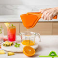 Orange juicer being used to extract juice into a measuring cup with a pink background.