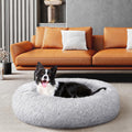 A black and white dog lies in a round, fluffy light gray pet bed placed in front of an orange leather sofa in a modern living room.