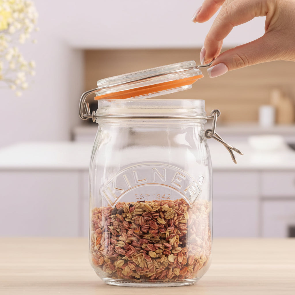 A clear glass jar with a clamp-lid seal and orange rubber gasket, filled with dried flowers or seed pods, sits on a light wood surface. A hand is lifting the lid, and a softly blurred indoor background is visible behind it.