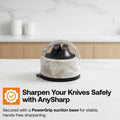 A close-up of a hands-free knife sharpener with a black PowerGrip suction base and a textured gray and white stone-like body. A finger is pressing the top handle, and garlic is blurred in the background on a wooden dish.