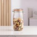 A clear Kilkin glass jar with a metal clamp lid and orange rubber seal, filled with assorted multicolored pasta, set on a light wooden table in a softly blurred kitchen background.