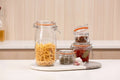 A clear glass clamp-lid jar filled with uncooked fusilli pasta sits on a white marble board, surrounded by smaller glass jars containing spices, garlic, and red chili peppers on a white countertop.