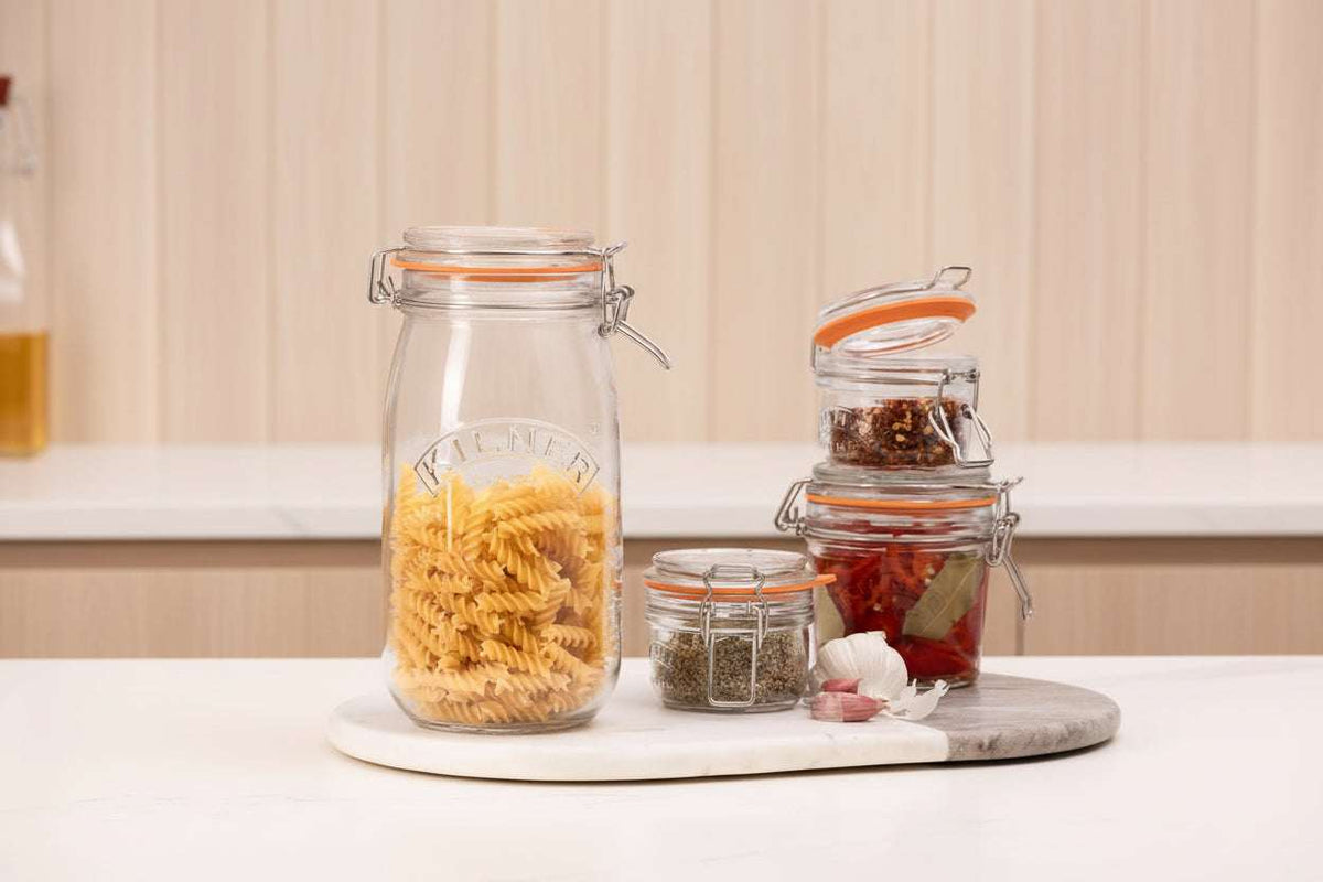 A clear glass clamp-lid jar filled with uncooked fusilli pasta sits on a white marble board, surrounded by smaller glass jars containing spices, garlic, and red chili peppers on a white countertop.