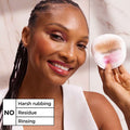 A smiling woman holds a makeup sponge with pink residue next to her face, with a text overlay that says 'NO Harsh rubbing Residue Rinsing' against a soft pink background.