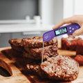 Digital thermometer being used to check the temperature of a cooked burger on a wooden cutting board.