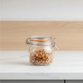 A clear glass clamp-lid jar with an orange seal, filled with almonds, on a light wooden surface against a soft lavender background.