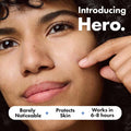 Close-up of a woman touching her cheek with a finger, with text reading 'Introducing Hero.' and a bottom banner that says 'Barely Noticeable + Protects Skin + Works in 6-8 hours.
