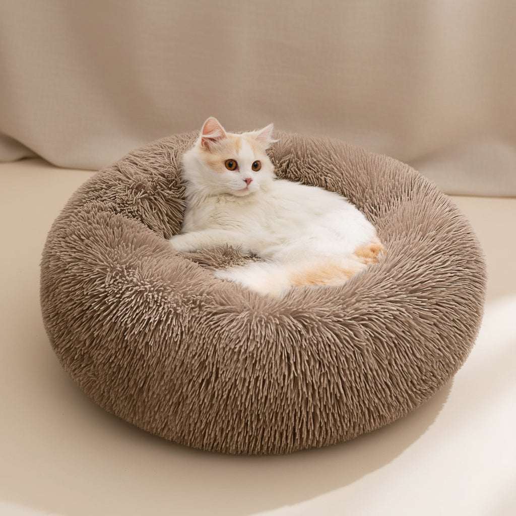 A fluffy white and orange cat rests inside a round, plush brown pet bed on a soft white faux-fur surface with sheer curtains in the background.
