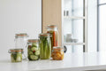 A set of clear glass clamp-lid jars with orange rubber seals, filled with various pickled vegetables and preserved foods, arranged on rustic wooden shelves next to fresh produce.