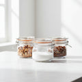 Three glass jars with metal clasps, containing various nuts and seeds, on a light surface.