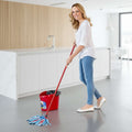 A woman in a white blouse and blue jeans mops a dark tiled kitchen floor with a red cleaning bucket and mop handle.