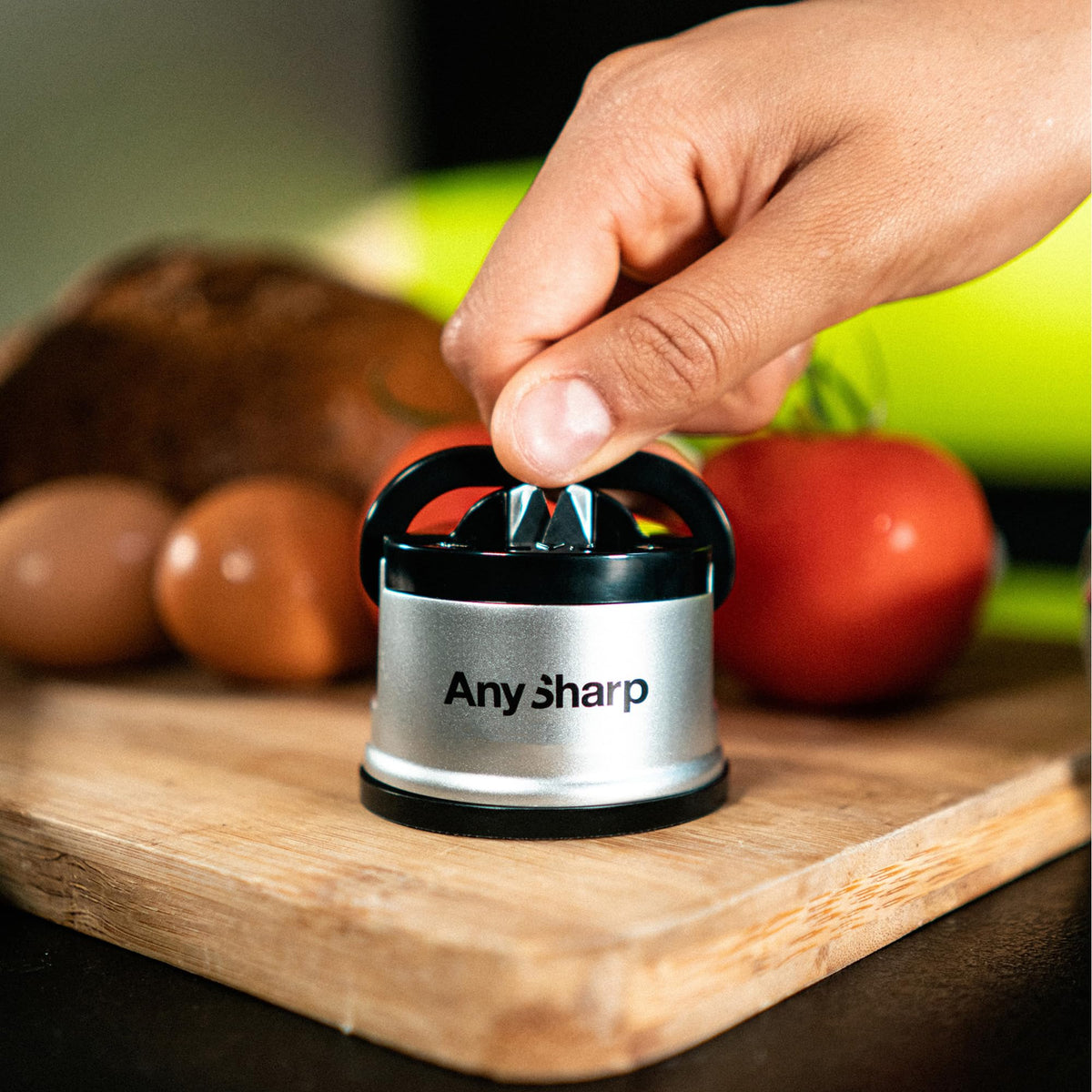 Hand using an 'Any Sharp' knife sharpener on a wooden cutting board with fruits in the background.