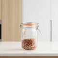 A Kilner glass jar with a metal clamp lid and orange rubber seal, filled with dried red and brown flakes, set on a light wooden surface with a softly blurred kitchen background.