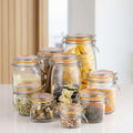 A set of clear glass clamp-lid jars with orange rubber seals, filled with various dry goods like pasta, rice, lentils, and spices, arranged on a white tablecloth against a light gray background.