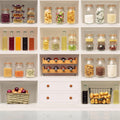 A 4x4 grid of white kitchen shelves filled with glass clamp-lid jars, bottles, baskets, and containers holding dried vegetables, spices, pasta, bread, and small bottles. No people or animals are present, and the setting is a clean interior shelving display.