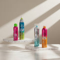 Four Bed Head hair styling aerosol cans displayed on gray concrete blocks against a textured gray wall. The cans are pink, green-to-blue gradient, silver-blue gradient, and pink-to-orange gradient, with bold text on the labels.