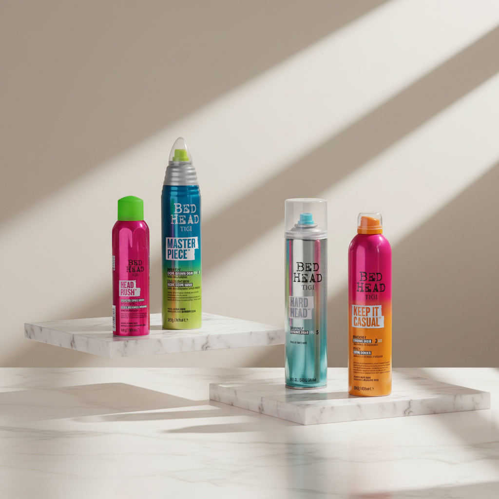 Four Bed Head hair styling aerosol cans displayed on gray concrete blocks against a textured gray wall. The cans are pink, green-to-blue gradient, silver-blue gradient, and pink-to-orange gradient, with bold text on the labels.