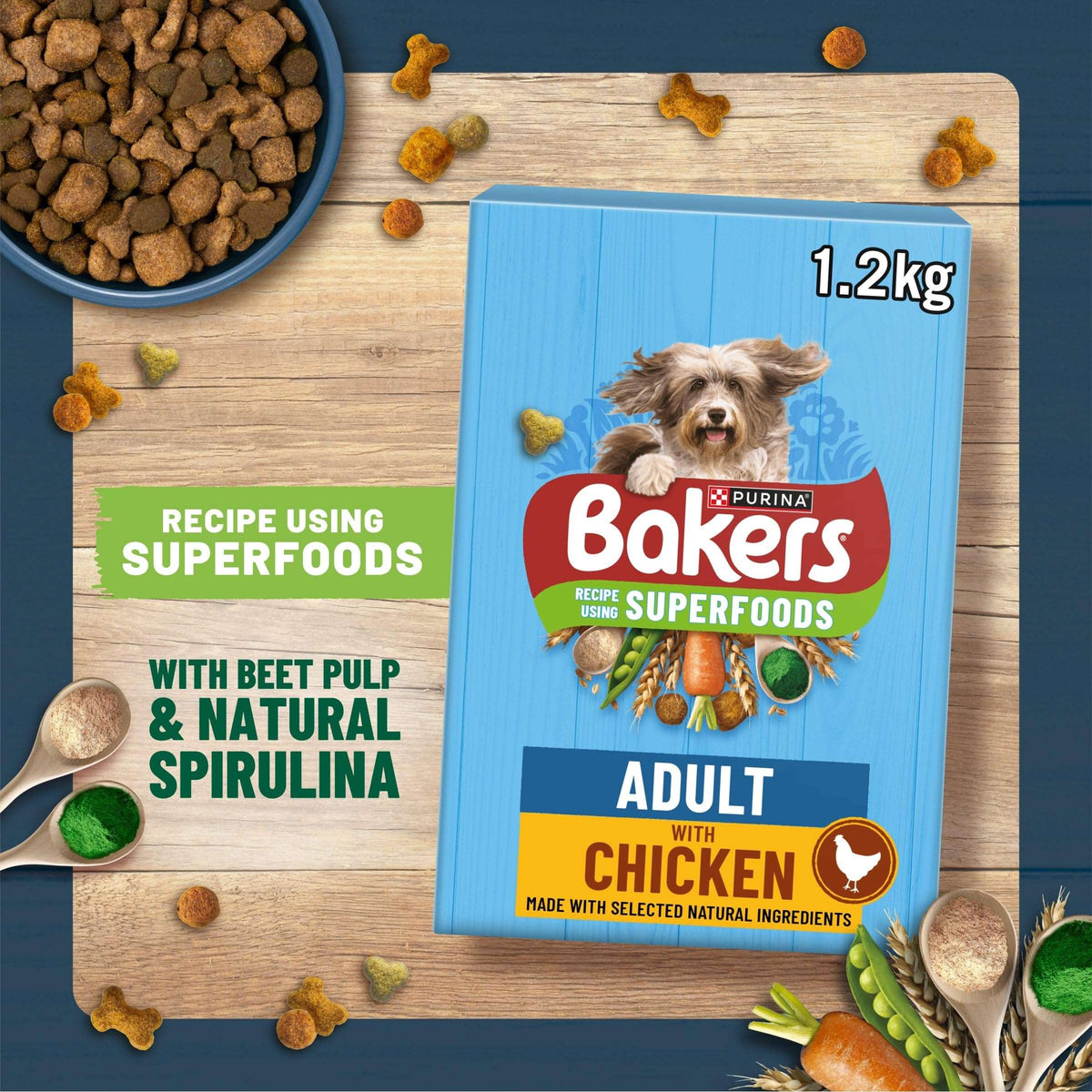A bag of Bakers Recipe 365 Superfoods Adult with Chicken dog food is displayed on a wooden board, surrounded by kibble, bowls, and ingredient props. The packaging is light blue with red, green, and yellow accents, and the text highlights the use of superfoods like beet pulp and spirulina.