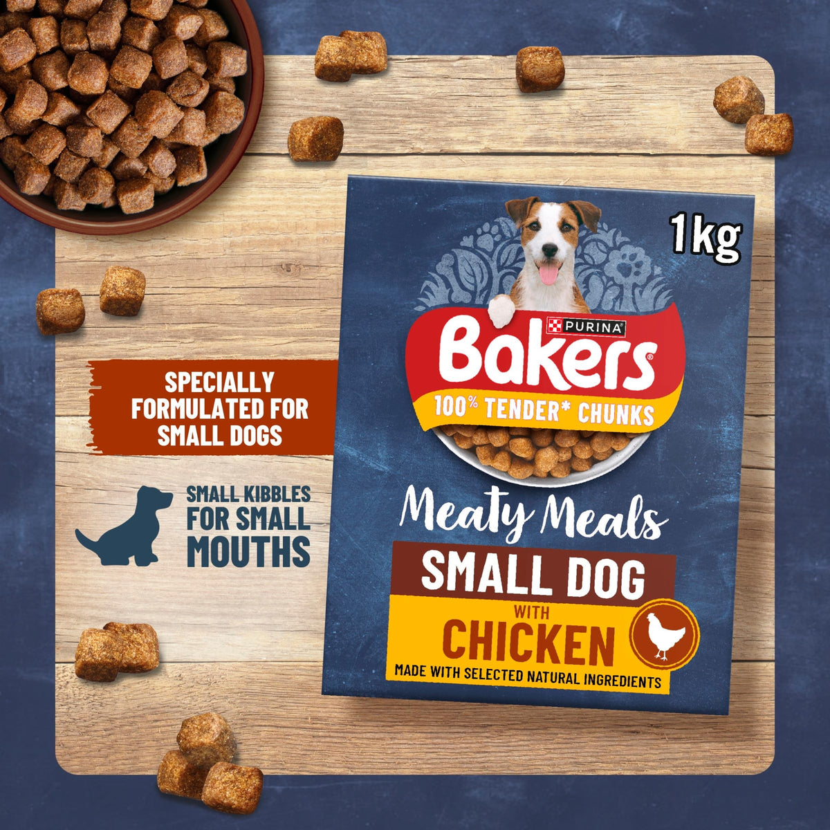 A 1kg package of Bakers Small Dog with Chicken dry dog food is displayed on a wooden board, surrounded by scattered kibble pieces and a bowl of dog food. The packaging is dark blue with red, yellow, and white branding, and includes a photo of a dog on the box.