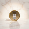 The bottom of a round gold-colored perfume tin held in a hand, showing printed text, a barcode, and a QR code on a white background.