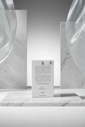 The back of a white rectangular perfume box with printed text, a signature, and an NFC logo, photographed against a neutral light gray background.