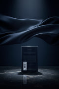 The back of a dark blue or black rectangular product box wrapped in clear plastic, with small white text and a barcode label near the bottom left, shown on a white background.